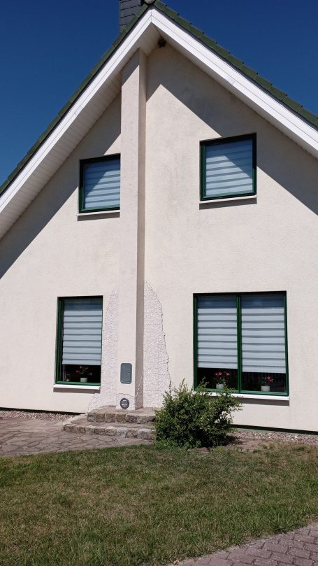 Helles Haus mit zwei Fenstern, grünen Fensterrahmen und einem kleinen Garten.
