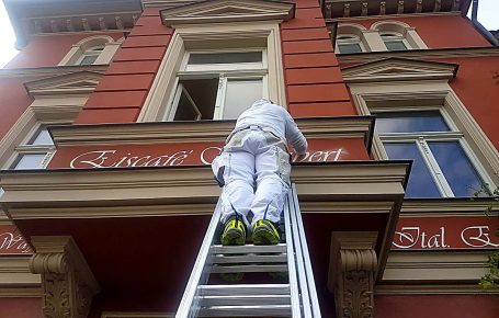 Ein Maler steigt eine Leiter hoch, um ein Fenster an einer roten Gebäudefassade zu streichen.