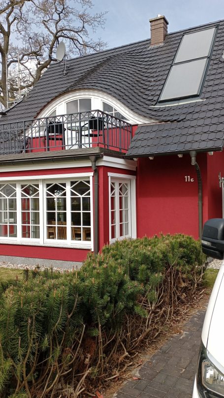 Rotes Haus mit einem Walmdach, großen Fenstern und Solarpanelen auf dem Dach.